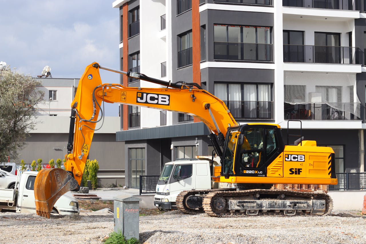 A yellow excavator operating at a city construction site in front of modern buildings.